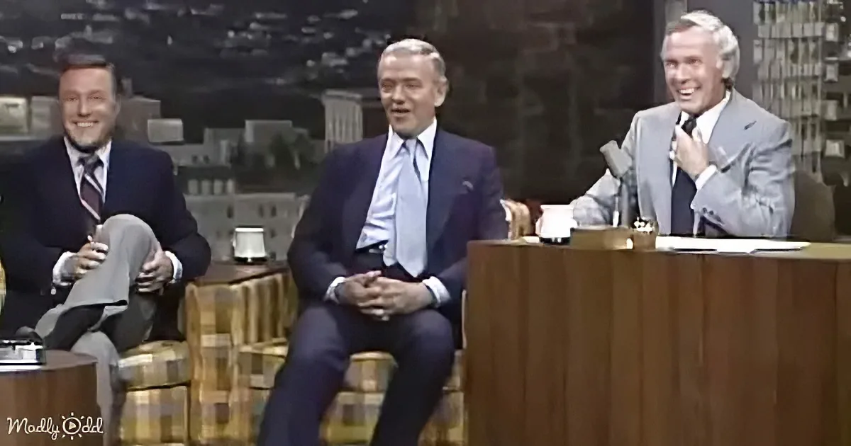 Three men in suits sit on a talk show set, one laughing, another smiling, and the third appearing relaxed.