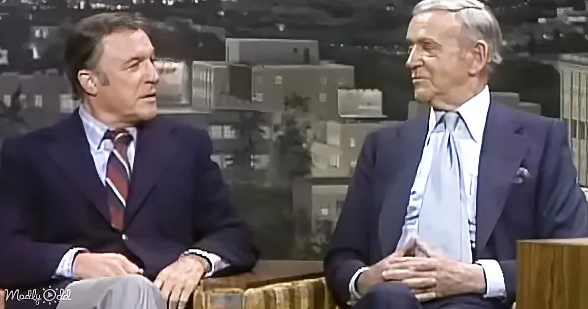 Two men in suits sit at a desk, one gesturing, in a studio setting with stacked boxes in the background.