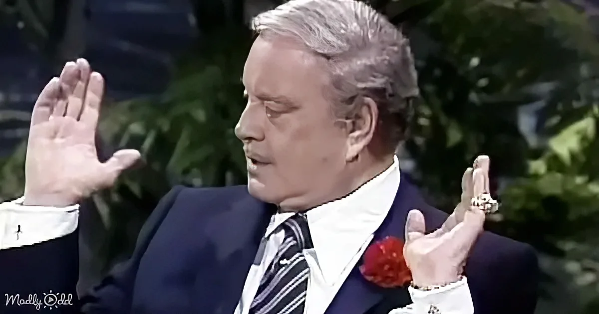 A man in a suit and tie, wearing a red boutonniere, gestures with his hands raised.