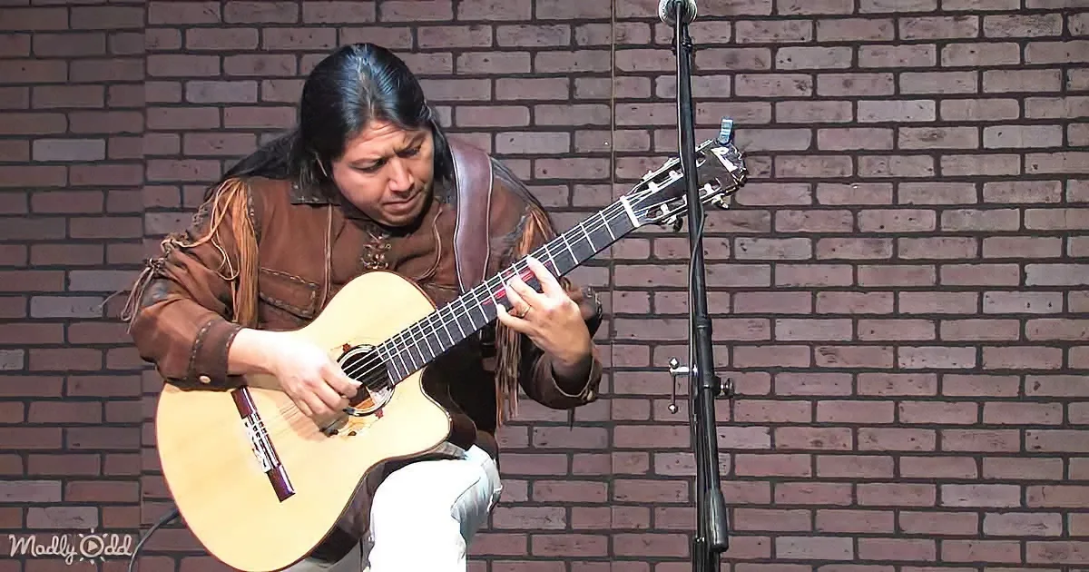 A person in a brown jacket plays an acoustic guitar against a brick wall.