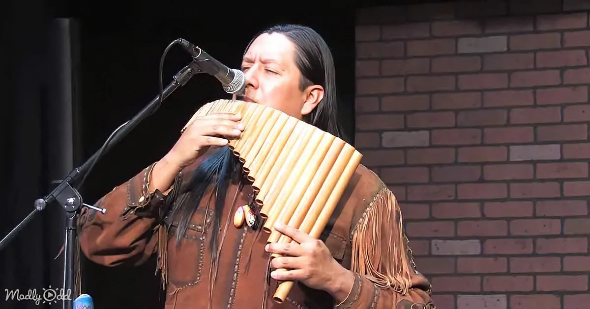 A person in traditional attire plays a pan flute on stage with a microphone and brick background.
