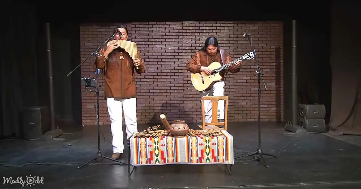 Two musicians perform on stage, one playing a pan flute and the other a guitar, with a colorful blanket and pottery on a...