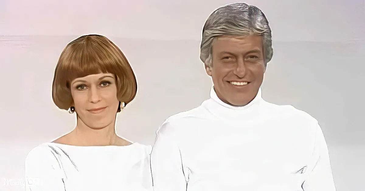 A man and woman in white outfits stand side by side, smiling, against a plain background.