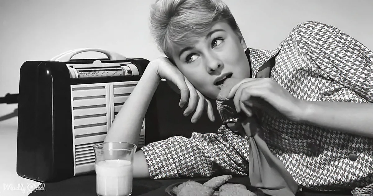A woman in a houndstooth dress leans on her hand, eating while looking up, with a milk glass and cookies nearby.