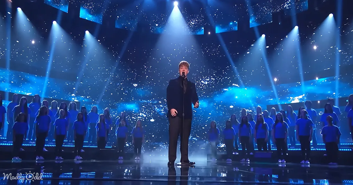 A man sings on stage with a choir in blue shirts, illuminated by blue lights and spotlights.