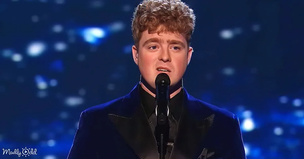 A person in a dark suit sings into a microphone against a blue, bokeh-lit background.