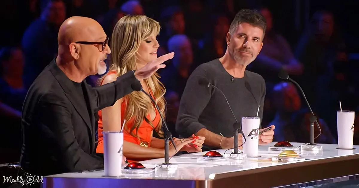 A man points while seated at a judging panel table with two other judges and a blurred audience in the background.