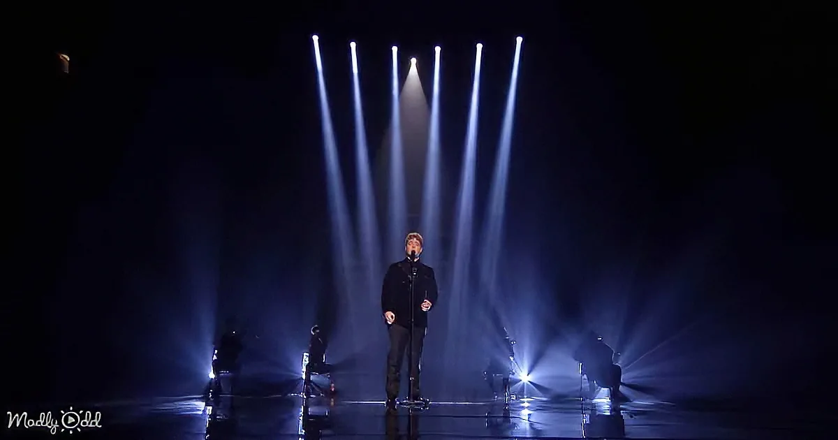 A man stands on stage under bright spotlights, surrounded by darkness, with a microphone in hand.