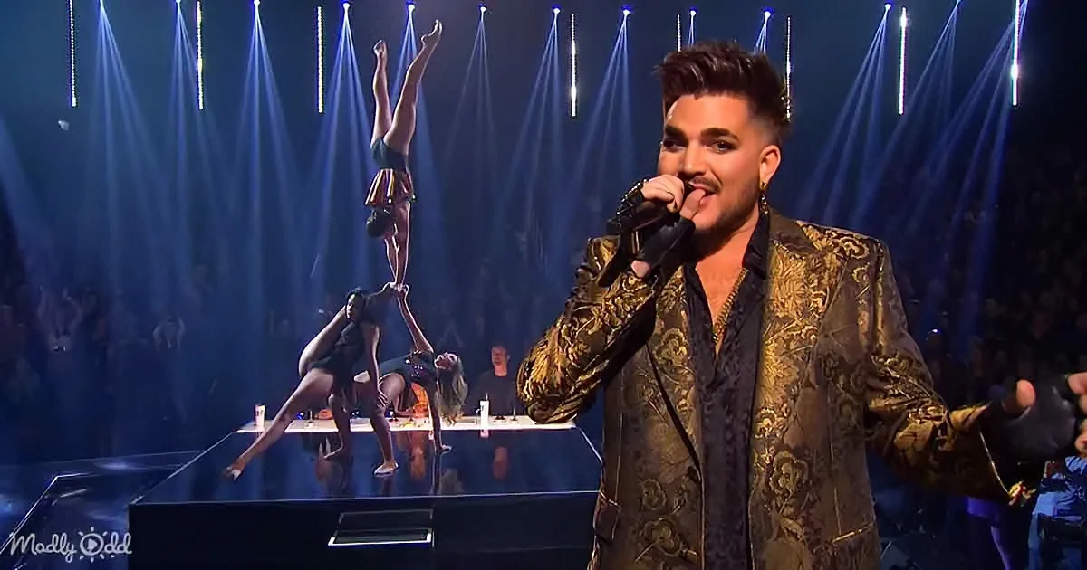 A performer in a gold jacket sings into a microphone on stage with acrobats in the background.