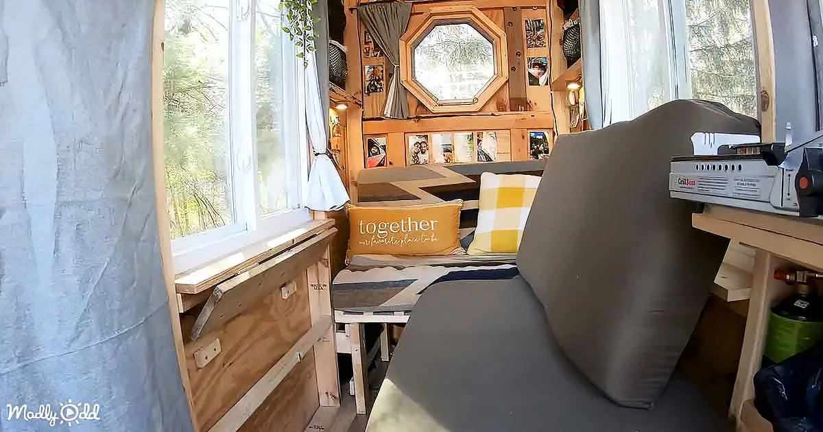 A cozy, compact living space with a wooden interior, a "together" pillow, and large windows showing greenery outside.