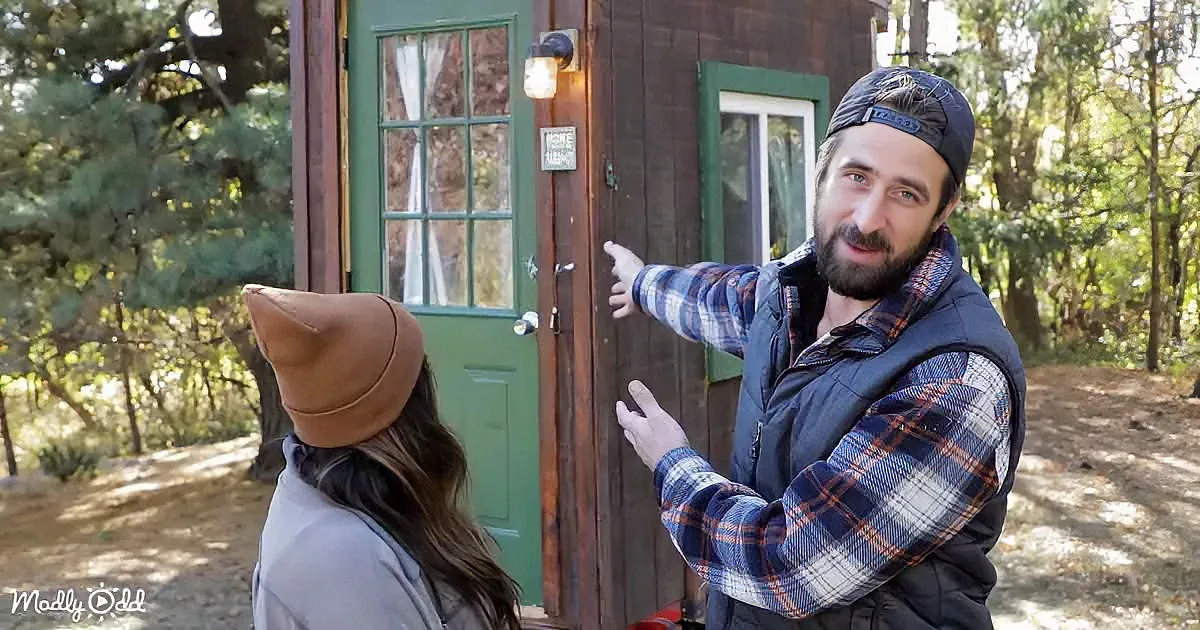 A man in a plaid shirt and hat points to a small cabin door while a woman in a brown hat looks on.