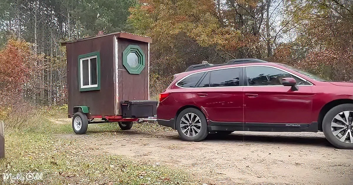 A red Subaru Outback towing a small brown trailer with a green window in a forested area.