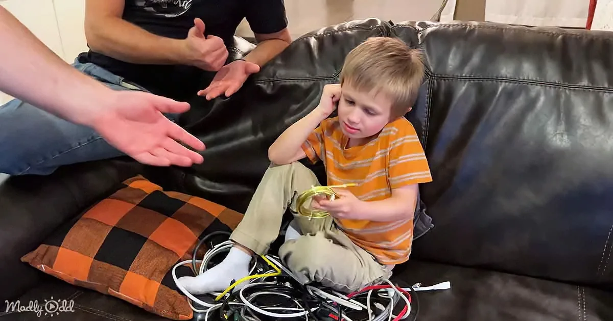 A young boy sits on a couch, holding a yellow cable amidst a pile of tangled wires.