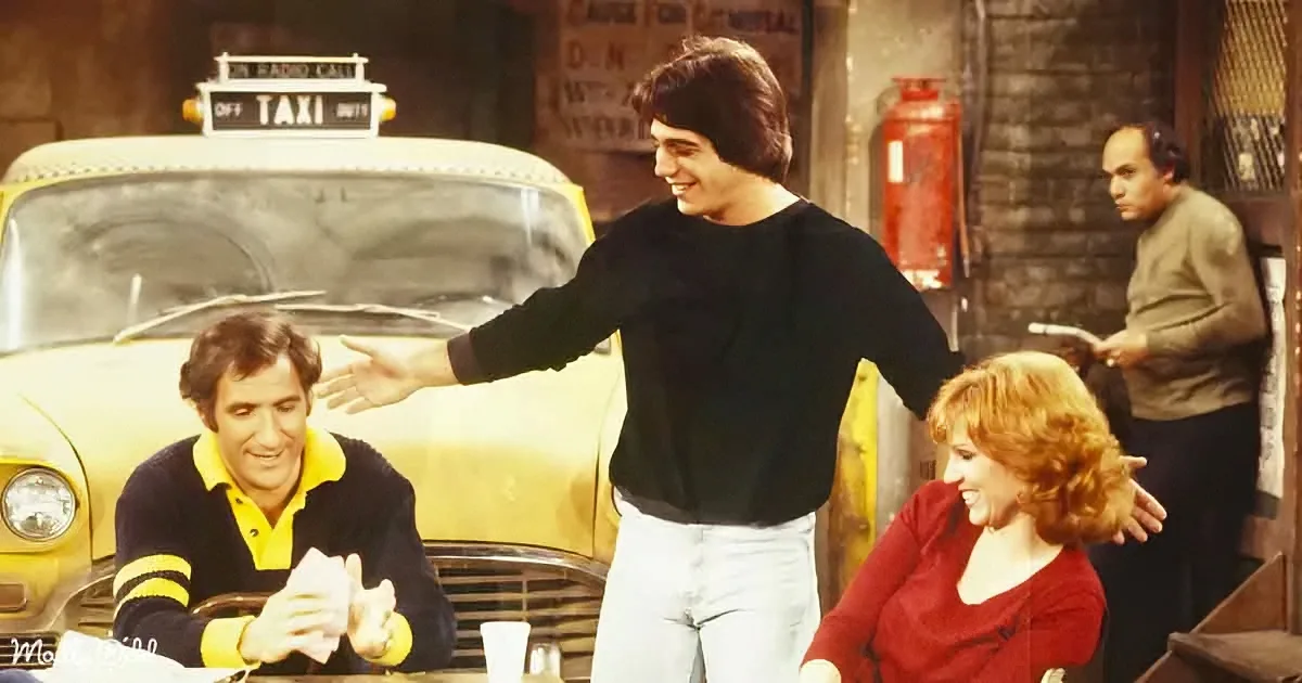 A man in a black sweater extends his hand, smiling, while a yellow taxi and two others look on in a casual setting.