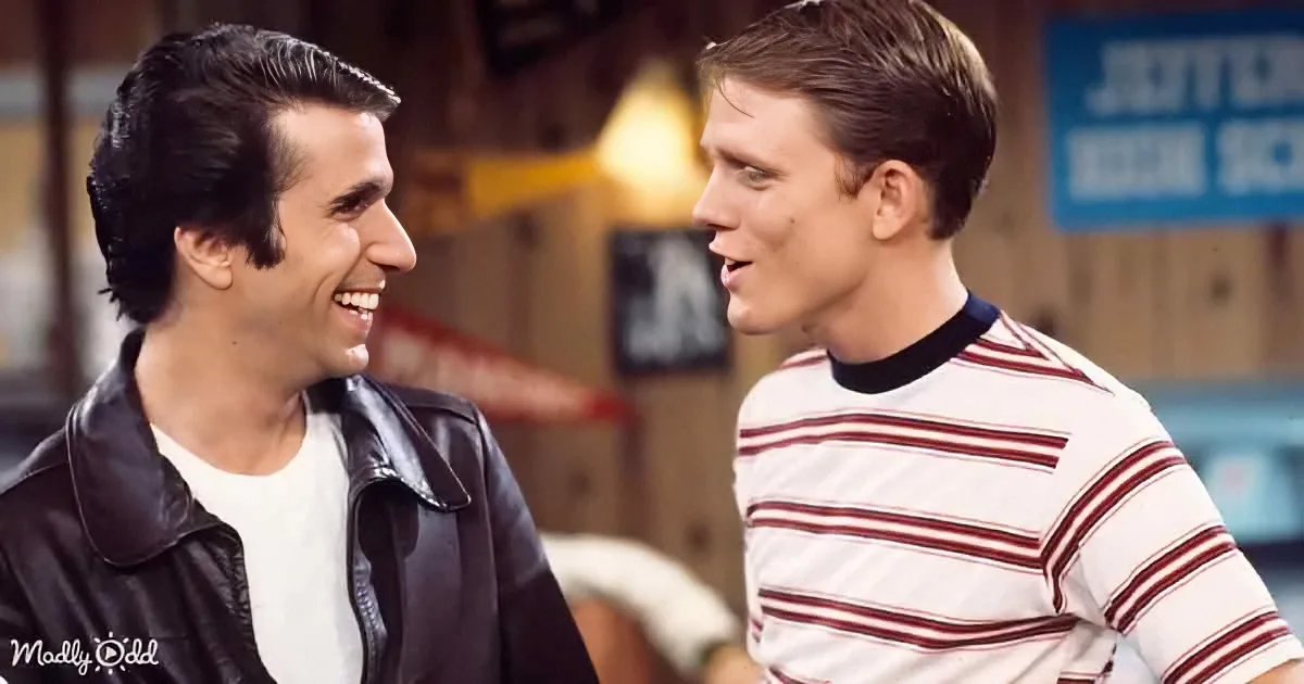 Two men in 1950s attire smile and talk, one in a leather jacket, the other in a striped shirt.
