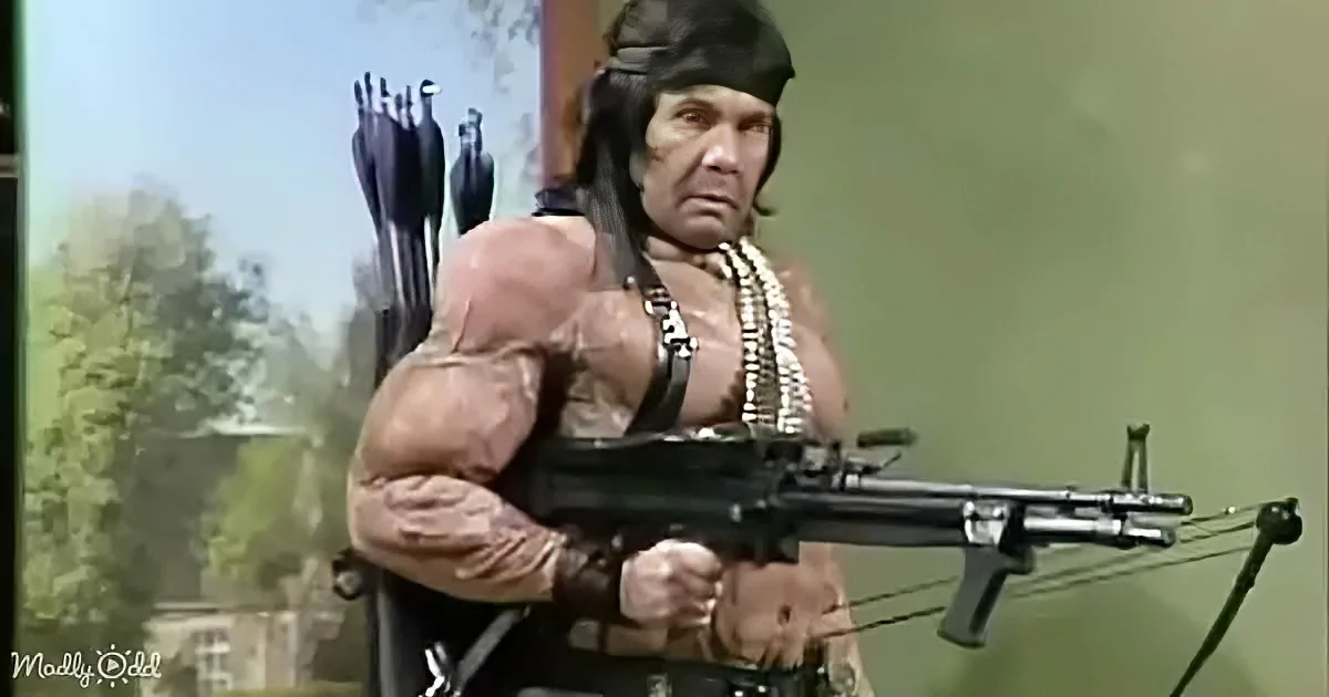 A muscular man in a costume holds a machine gun and arrows, wearing a headdress and beads.