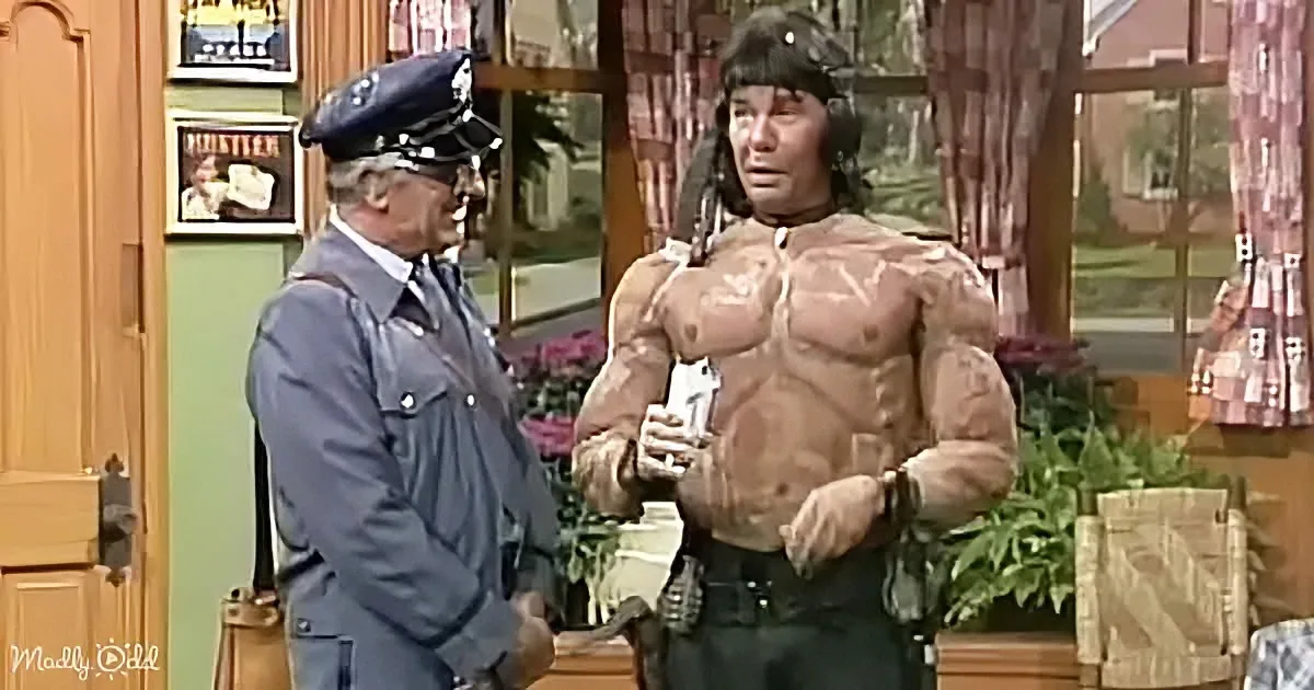A muscular man in a police uniform stands next to a man in a blue uniform, both in a room with a window and plants.
