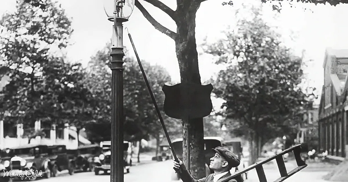A man climbs a ladder to adjust a street lamp in a tree-lined street with vintage cars.