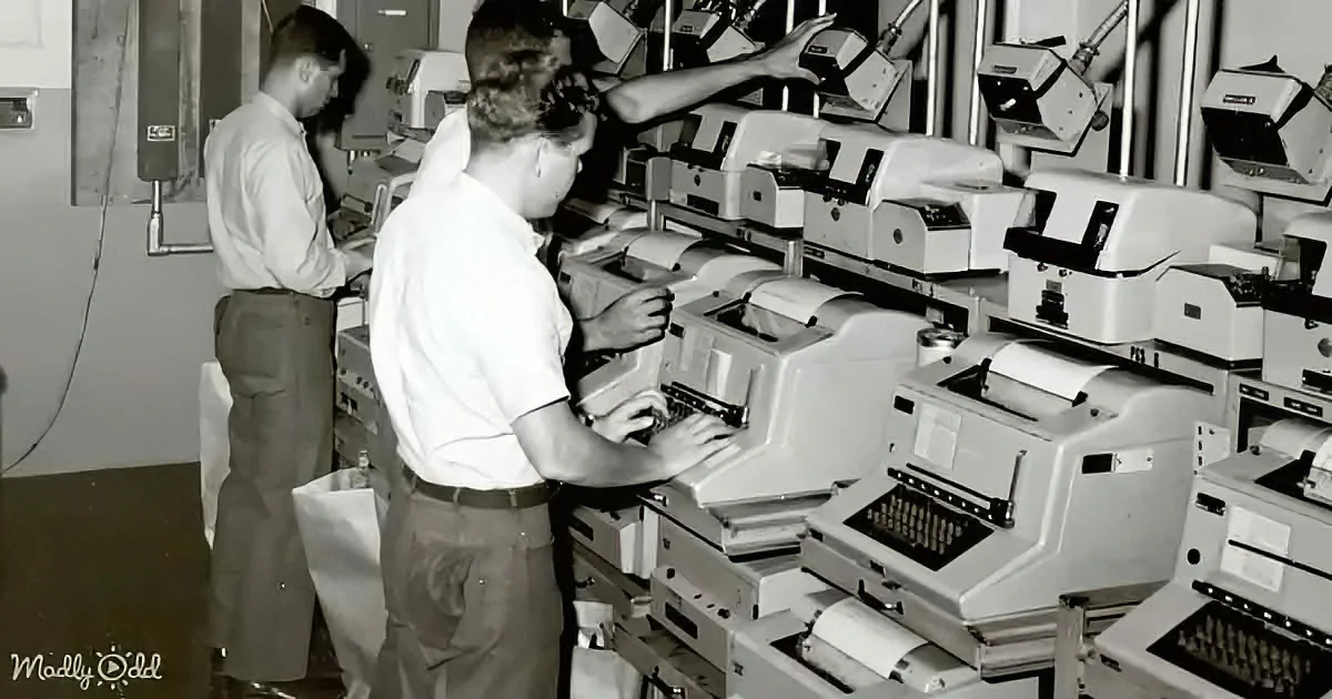 A man operates a large, old-fashioned computer terminal surrounded by similar machines in a room.