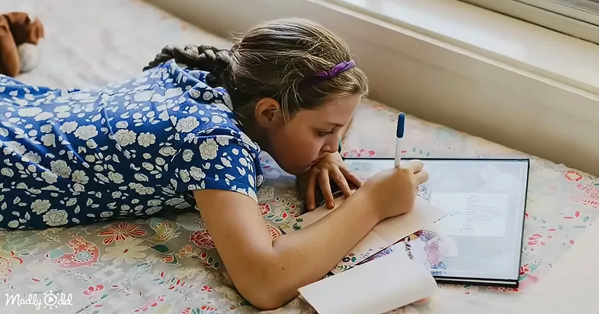 A girl in a floral dress lies on a patterned surface, writing in a notebook with a pen.