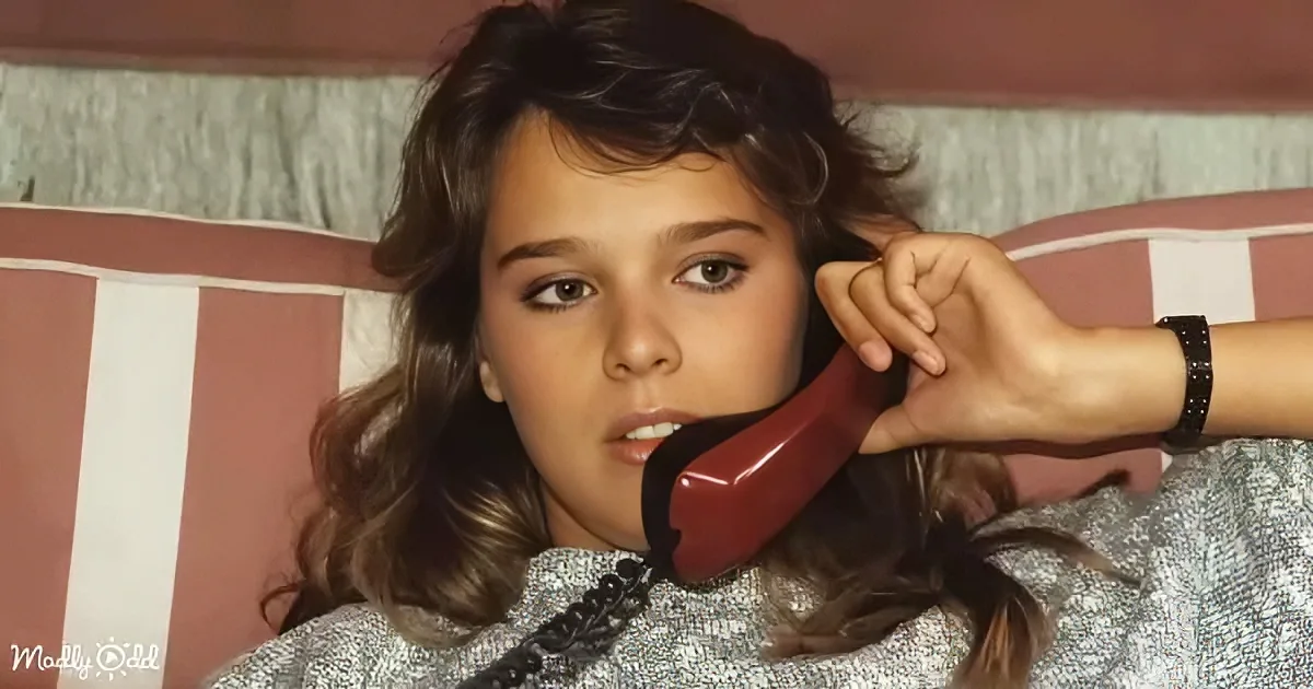 A person with long, wavy hair is holding a red telephone receiver to their ear while lying on a striped couch.