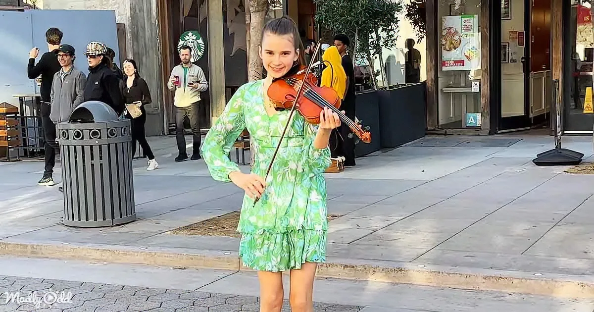 A girl in a green floral dress holds a violin on a sidewalk, smiling, with people and a Starbucks in the background.