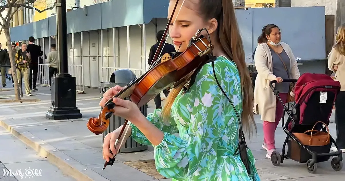 A woman in a floral dress plays the violin on a city sidewalk, surrounded by pedestrians and a stroller.