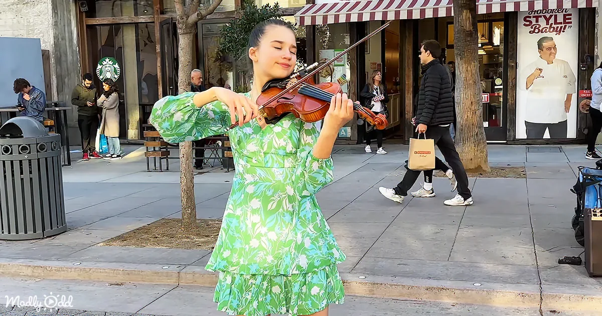 A woman in a green floral dress plays the violin on a city sidewalk, surrounded by pedestrians and storefronts.