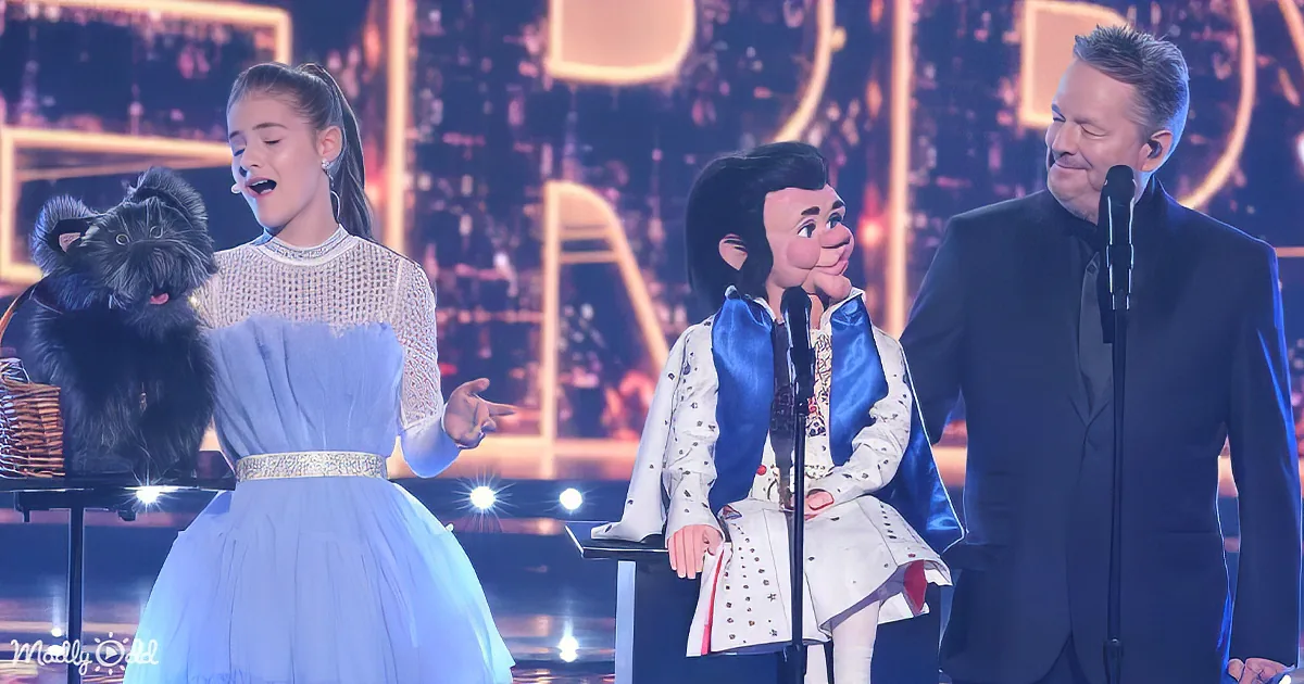 A young girl in a blue dress sings on stage with a puppet and a man in a suit.