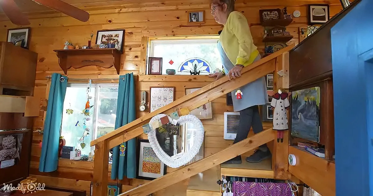 A woman descends a wooden staircase in a cozy, rustic cabin adorned with art and personal items.