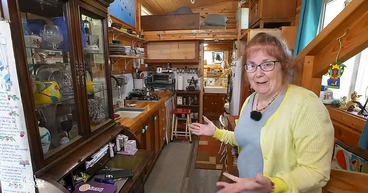 A woman in a yellow cardigan stands in a cozy, cluttered kitchen with wooden cabinets and a colorful rug.