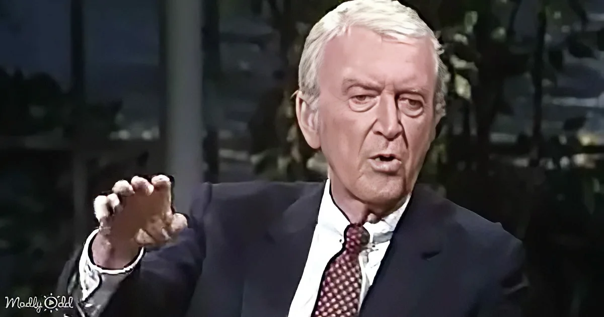 An older man in a suit gestures with his right hand while speaking.