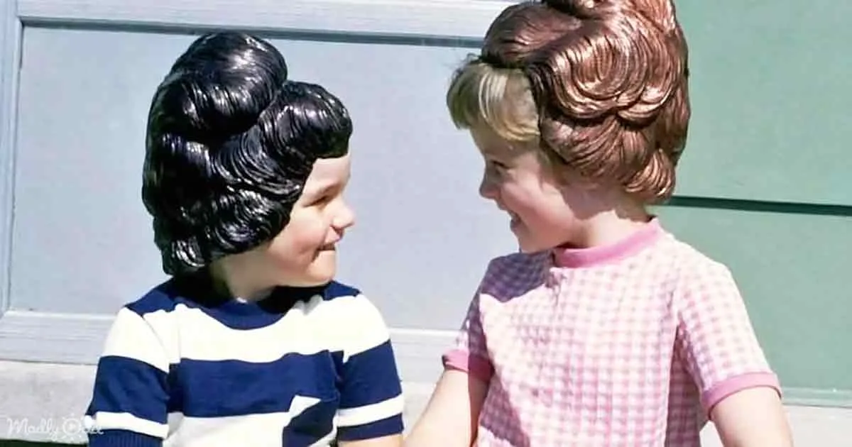 Two children in vintage outfits and wigs, one in a striped shirt, the other in a checkered dress, facing each other.