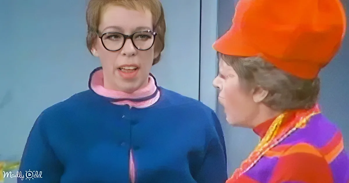 A woman in a blue jacket and glasses stands next to another in a colorful outfit and orange hat.