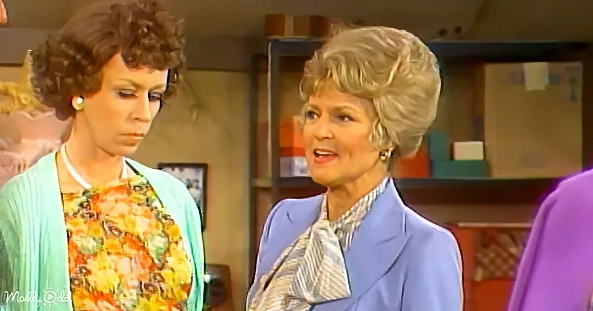 A woman in a blue suit and patterned tie stands next to another woman in a floral dress, both in a room with shelves.
