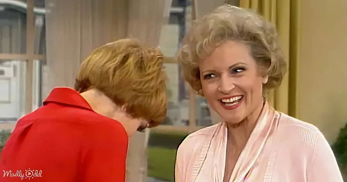 A woman in a pink blouse smiles while another in a red jacket looks down, both in a room with a window and curtains.