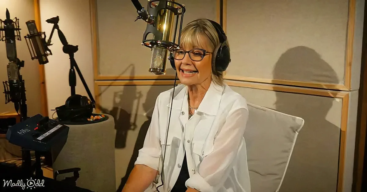 A woman in a recording studio smiles while wearing headphones and glasses, surrounded by microphones and soundproofing...