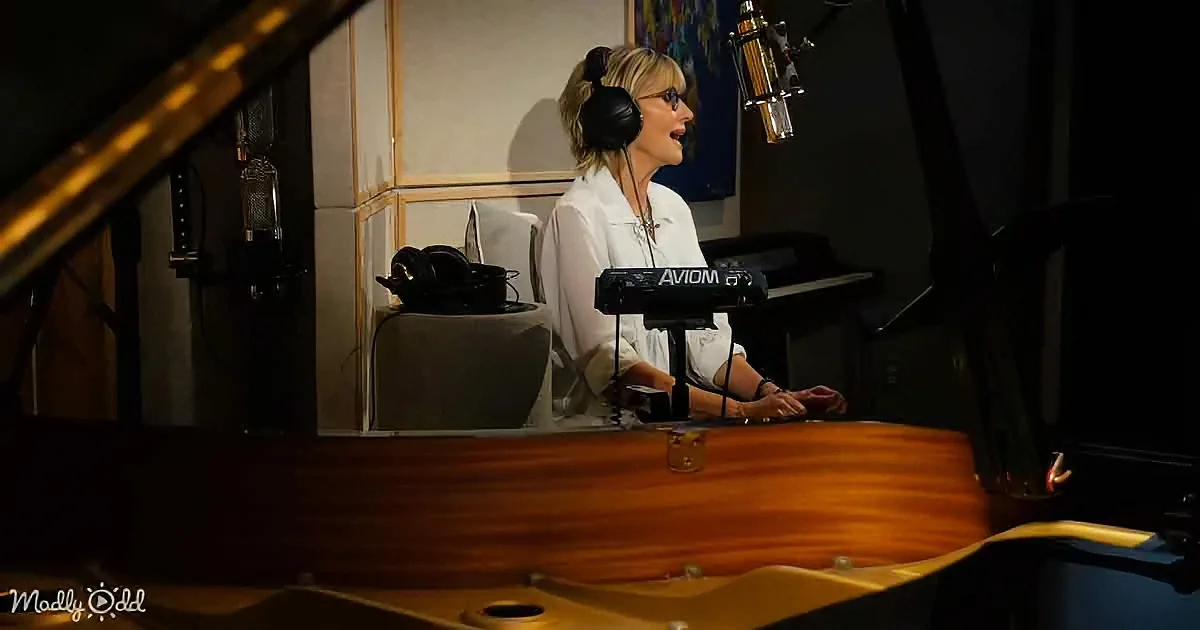 A woman sings into a microphone in a recording studio, wearing headphones and a white shirt.