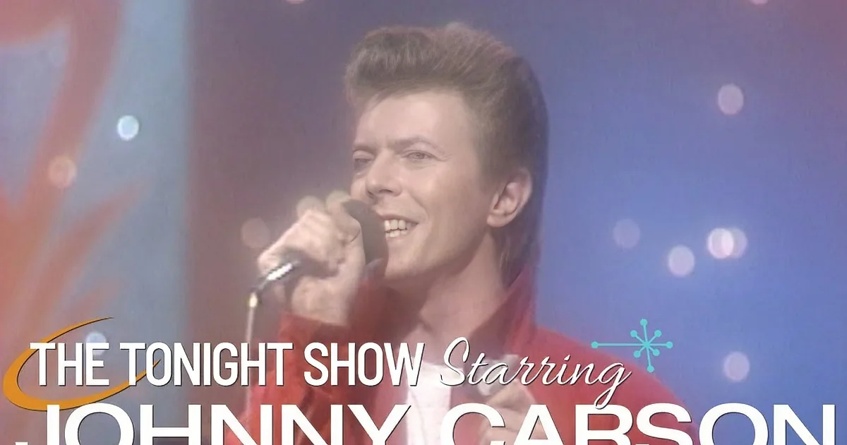 A man in a red jacket smiles while singing into a microphone, with blurred lights in the background.