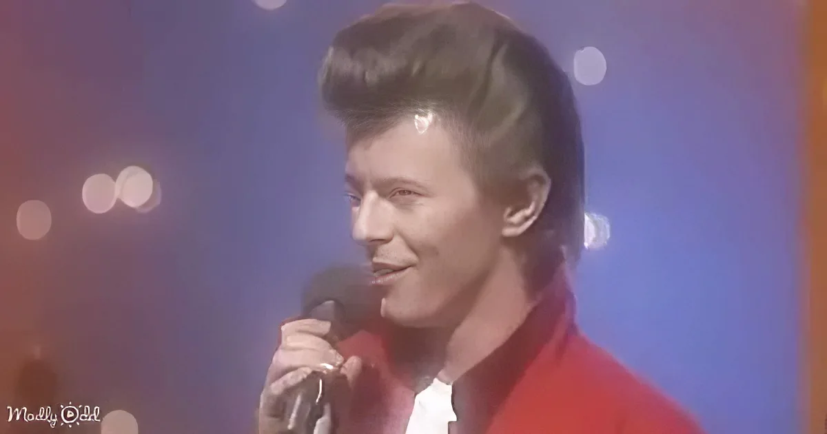 A person in a red outfit holds a microphone, smiling, with a blurred background of lights.