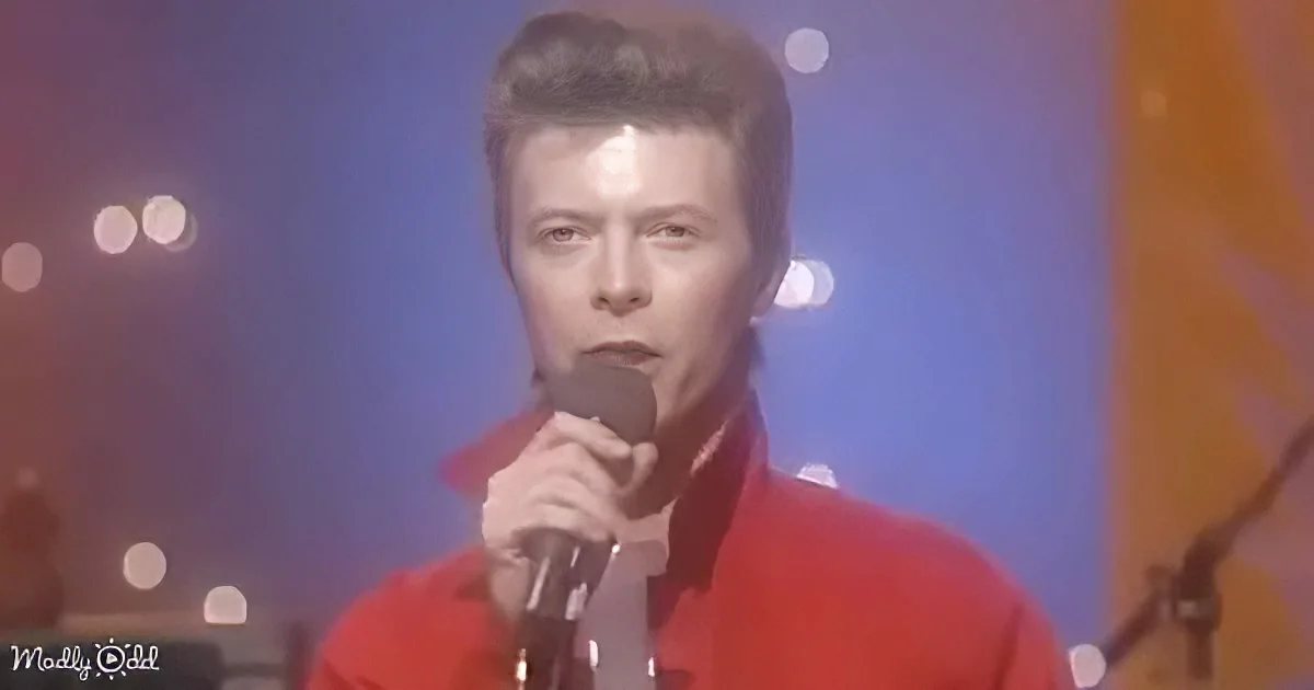 A person in a red outfit holds a microphone, singing on stage with blurred lights in the background.