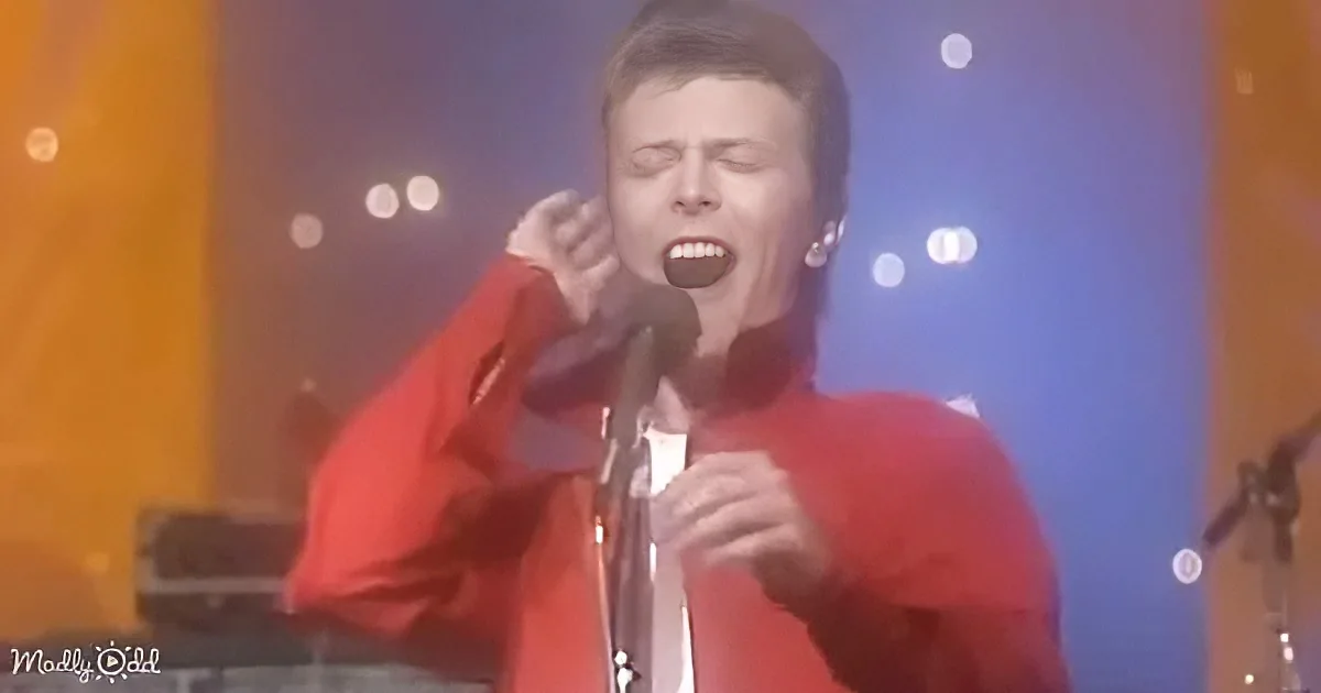 A person in a red outfit sings into a microphone on stage with blurred lights in the background.