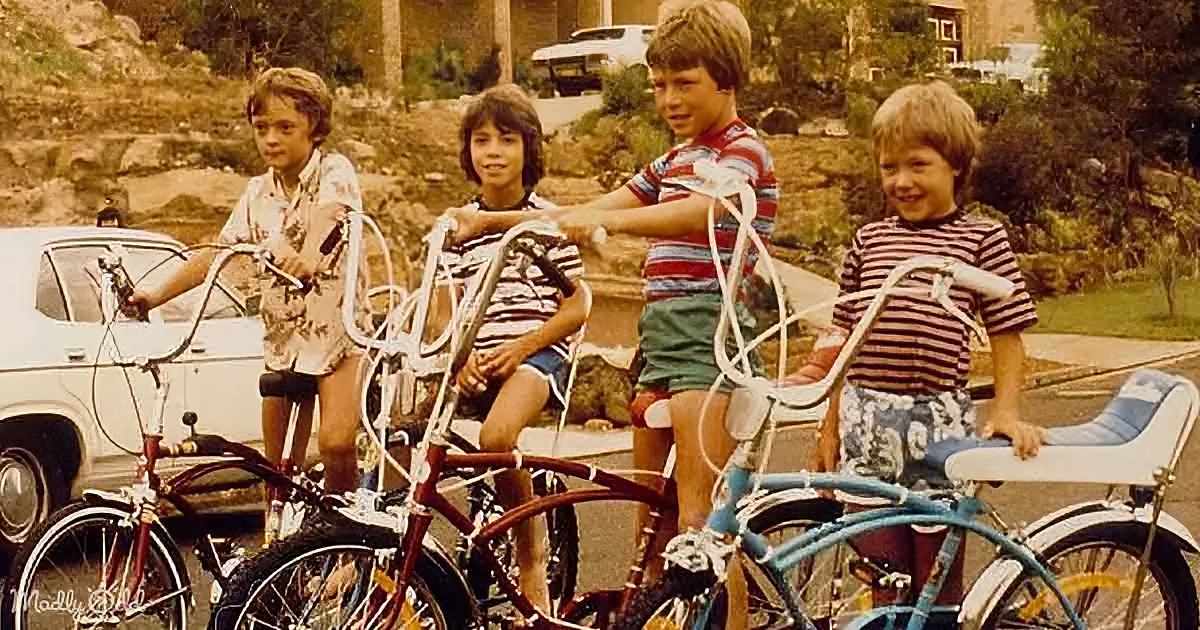 Four young boys with bicycles smile in a suburban driveway, vintage cars and houses in the background.