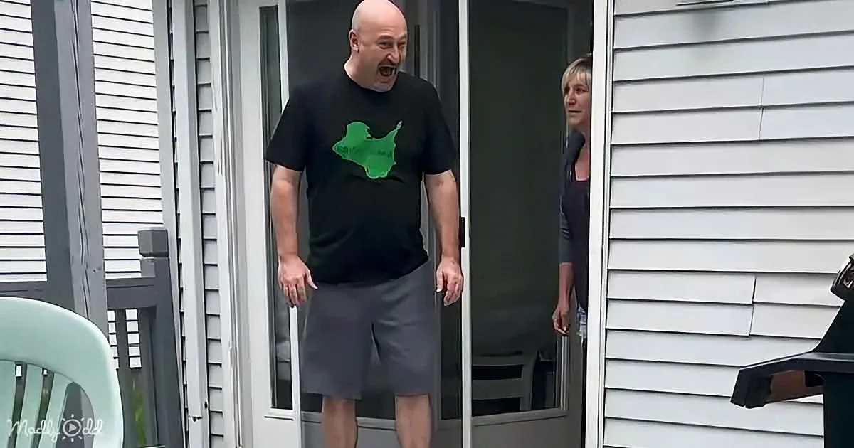 A man in a black shirt with a green graphic stands in a doorway, laughing, while a woman looks on.