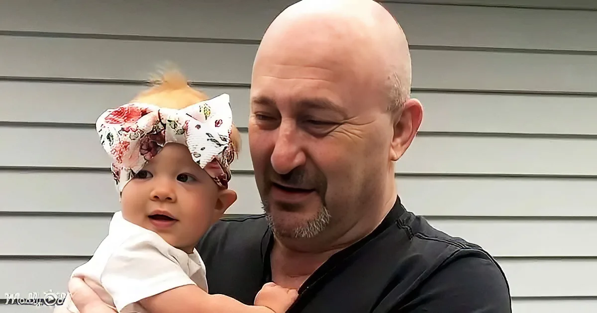 A man holds a baby with a floral bow, both smiling, against a gray siding background.
