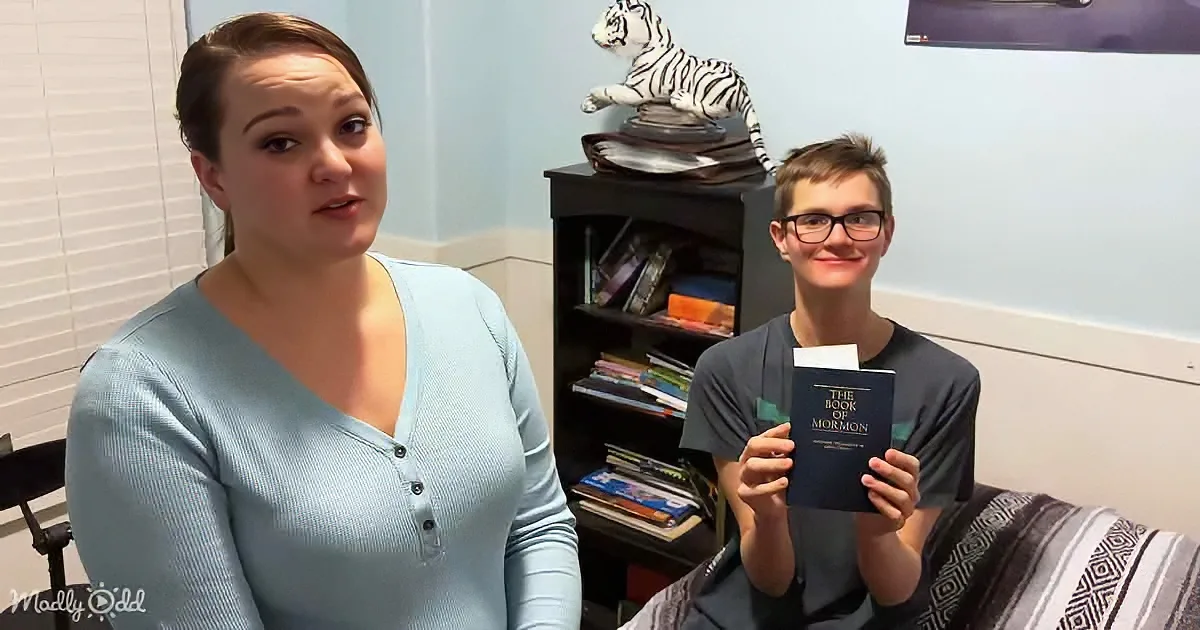 A woman in a blue shirt sits beside a boy holding "The Book of Mormon," with a bookshelf and tiger statue in the background.