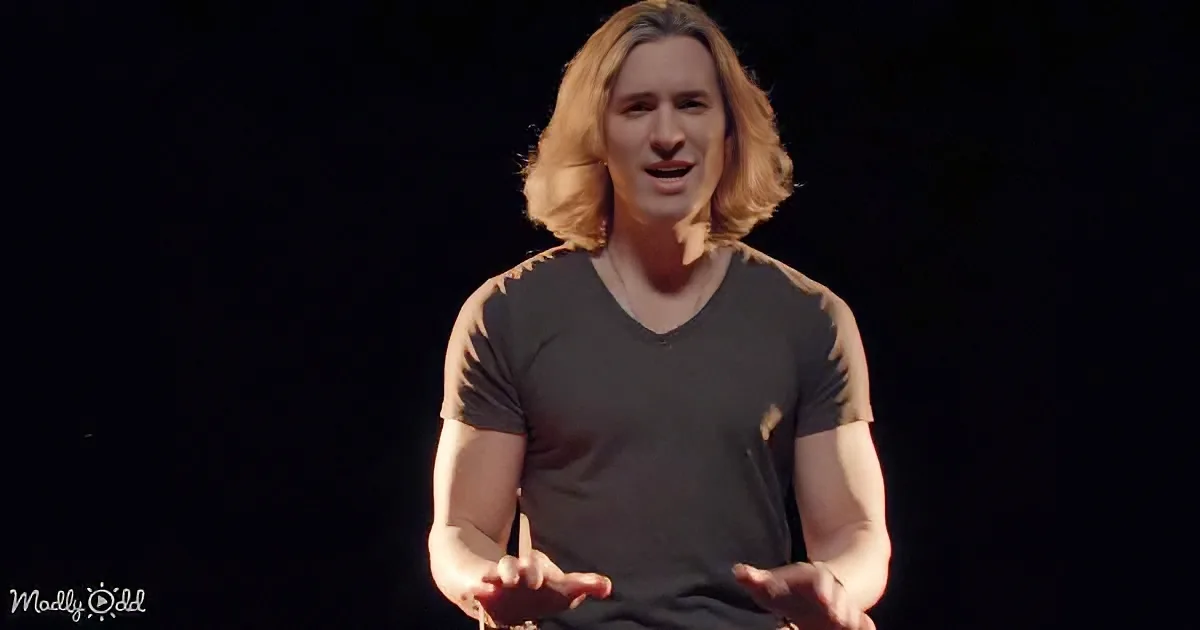 A person with long hair, wearing a dark V-neck shirt, stands against a black background, gesturing with their hands.