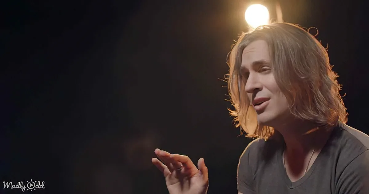 A person with long, wavy hair gestures while speaking, illuminated by a light source behind them.