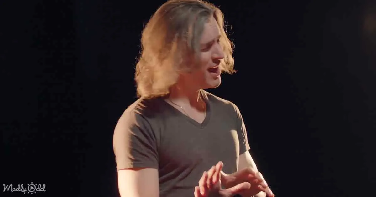 A person with shoulder-length hair, wearing a dark t-shirt, is gesturing with their hands against a dark background.