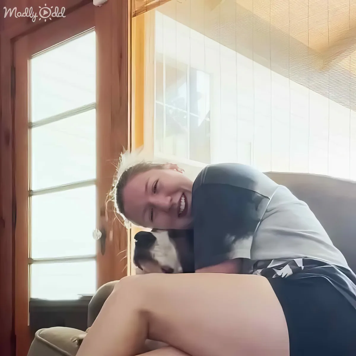 A person is hugging a dog on a couch, both smiling, with a window and door in the background.